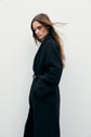 A person with long brown hair wearing a chic à la française belted dark manteau long stands against a plain white background, looking down with wind blowing through their hair.