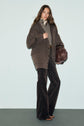 A woman stands against a plain background wearing a stylish manteau mi-long in brown, layered over a gray turtleneck, with patterned wide-leg pants and brown shoes, holding a large round brown bag.