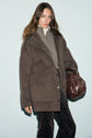 A woman wearing a brown oversized manteau laine, gray zip-up sweater, white turtleneck, and patterned pants holds a brown leather bag. She stands against a plain light blue background.