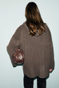A person with long brown hair, seen from behind, wears an oversized manteau laine in brown and carries a brown leather bag, standing against a light blue background.