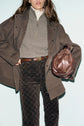 Woman wearing a brown manteau mi-long, gray zip-up sweater, patterned brown pants, and holding a brown leather purse. Her face is partially out of frame. Background is light blue.