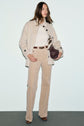 A woman stands against a pale background, wearing a manteau écru—a cream jacket—paired with a white sweater, tan corduroy pants, brown belt, brown boots, and holding a dark textured handbag.