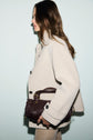 A woman with long brown hair wearing a manteau écru stands in profile, holding a dark brown woven handbag against a plain light background.
