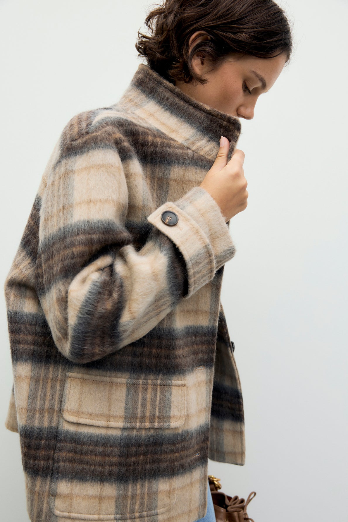 A person in a beige and brown manteau à carreaux with large buttons holds the collar up, looking down against a plain light background.