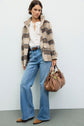 Woman standing against a light wall, wearing a manteau à carreaux—a plaid jacket—in soft wool mohair, paired with a white blouse, wide-leg jeans, brown shoes, and holding a tan leather handbag. She has short wavy hair and a calm expression.