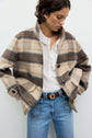 A woman wearing a beige and brown manteau à carreaux over a white ruffled blouse and blue jeans stands with hands in her pockets, looking down.