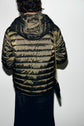 Person facing away, wearing a shiny, puffy, olive-green doudoune légère with a hood. They have dark, wavy hair and are carrying a small black bag with a gold chain strap over their right shoulder.