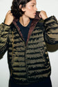 A person wearing a shiny olive green doudoune légère with a brown lining looks to the side while adjusting the hood.