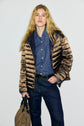 A person with long blonde hair wears a doudoune ultra légère metallic brown puffer jacket over a blue button-up shirt and dark jeans, holding a tan bag, standing against a plain light background.