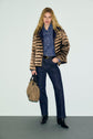 A person with long blonde hair wears a shiny brown doudoune femme puffer jacket, blue denim shirt, dark jeans, and black boots, holding a tan bag, standing against a plain light background.