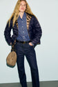 Person wearing a doudoune ultra légère dark blue jacket over a denim shirt and jeans stands with one hand in their pocket, holding a beige bag in the other hand, against a light background.