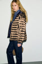 A person with long blonde hair wears a shiny bronze doudoune femme puffer jacket over a blue shirt and dark jeans, standing against a plain white wall.