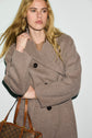 A person with long blonde hair wearing a manteau long laine brown coat holds the collar and carries a patterned brown handbag.