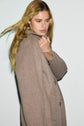 A person with long blonde hair wearing a light brown manteau laine femme looks down and slightly to the side, standing against a plain white background.