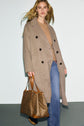 A woman wearing a manteau long laine in beige, paired with a tan sweater and blue jeans, holds a large brown patterned handbag with leather straps as she walks against a plain light background.
