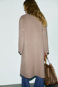A person with long, wavy hair wears a manteau long laine beige and blue jeans, standing with their back to the camera. They hold a brown patterned handbag in their right hand.