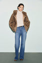 A person stands against a plain background wearing a brown manteau fausse fourrure over a beige turtleneck sweater, blue high-waisted jeans, and brown shoes.