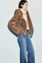 A person wearing a light brown manteau fausse fourrure over a white turtleneck sweater paired with high-waisted blue jeans stands against a pale background, looking to the left.