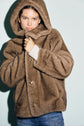 A person wearing a hooded, brown manteau fausse fourrure with large buttons stands against a light background, holding the manteau capuche closed at the neck and looking directly at the camera.