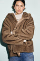 A person wearing a brown manteau fausse fourrure over a white turtleneck sweater and blue jeans stands against a light blue background, looking slightly to the side with arms crossed.