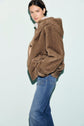 Person wearing a brown manteau court en fausse fourrure avec capuche and blue jeans stands against a light blue background, looking at the camera with a neutral expression.