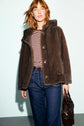 A person with red hair wears a brown manteau fausse fourrure, a red and white striped shirt, and blue jeans while holding a dark handbag against a light background.