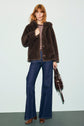 Woman with red hair wearing a manteau fausse fourrure in brown, a striped shirt, flared blue jeans, and brown boots, holding a brown handbag, stands against a pale background.