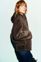 A person with reddish hair wearing a brown manteau à capuche en fausse fourrure and dark pants stands against a pale blue background, looking at the camera.