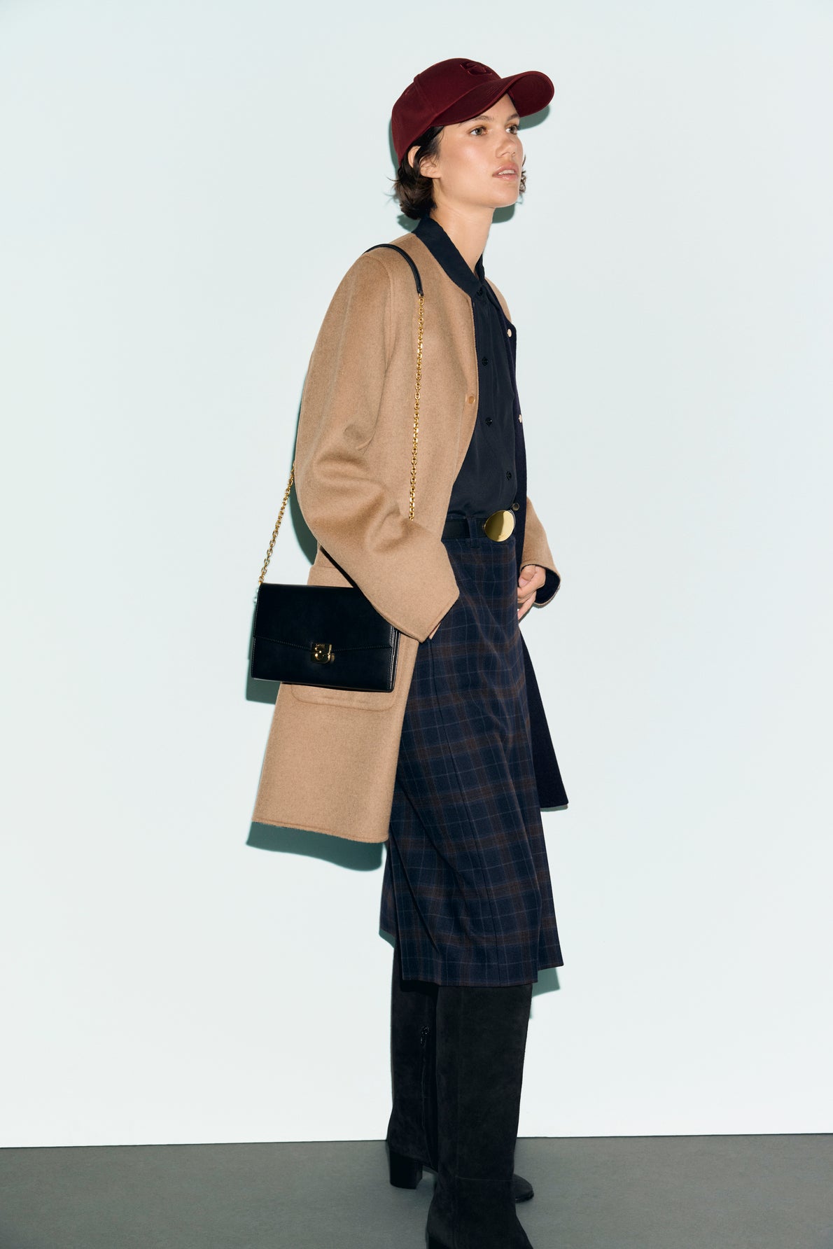 A person wearing a red cap, a manteau en laine with boutons pression dorés, black shirt, plaid skirt, tall black boots, and carrying a black shoulder bag with a gold chain stands against a light background.