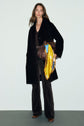 Woman stands against a plain background wearing a manteau mi-long in black, paired with patterned black pants and brown shoes. She holds a brown bag adorned with a bright yellow and blue scarf tied to it.