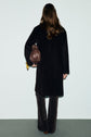 A person with long brown hair, wearing a manteau mi-long femme in black and patterned dark pants, stands facing away, holding a brown handbag in their left hand.