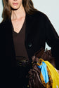 A woman wearing a black manteau mi-long and a brown top holds a brown handbag decorated with yellow and blue scarves. The image shows her from the shoulders to the waist, with part of her face visible.