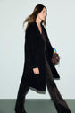 Woman walking in profile, wearing a manteau mi-long noir, dark patterned pants, and holding a brown handbag with a blue accessory, against a plain light background.