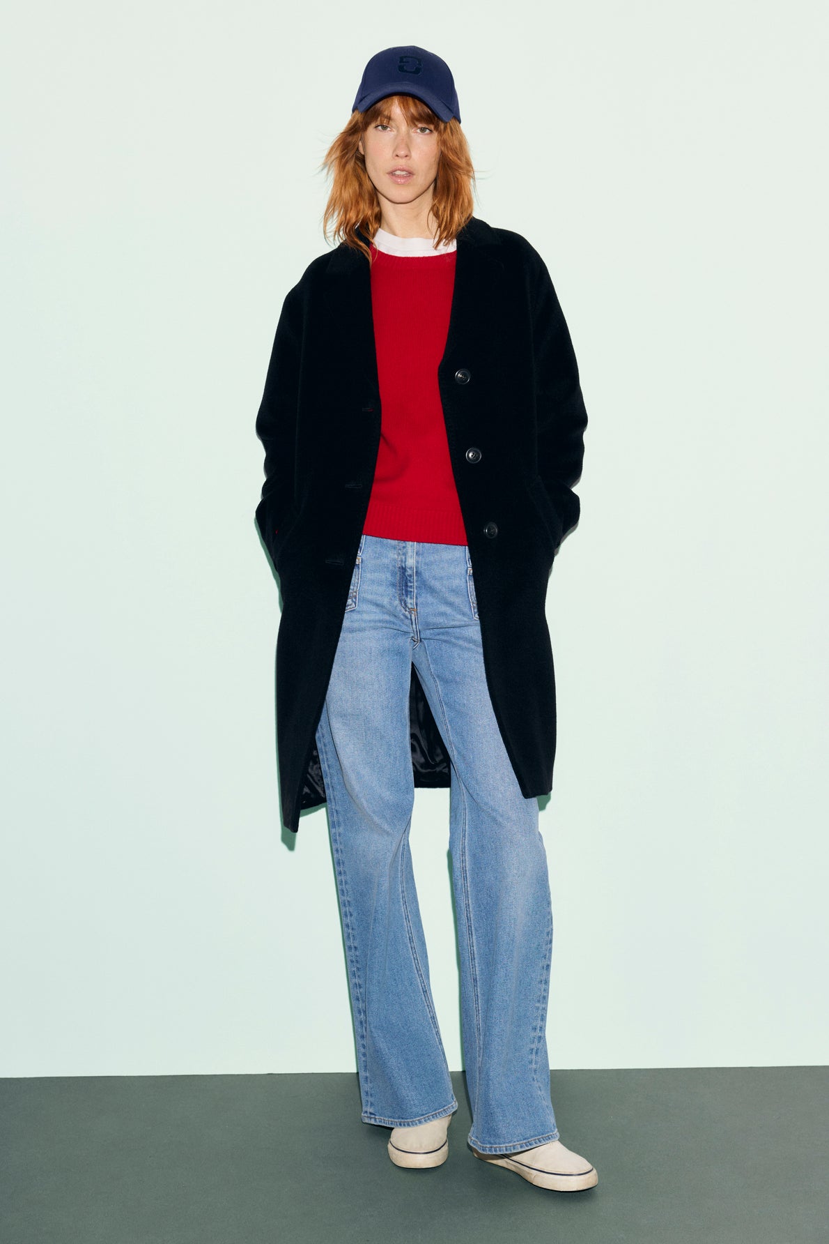 Woman wearing a navy cap, long manteau noir en laine angora, red sweater, light blue flared jeans, and beige shoes stands against a plain pale green background, looking at the camera with hands in her coat pockets.
