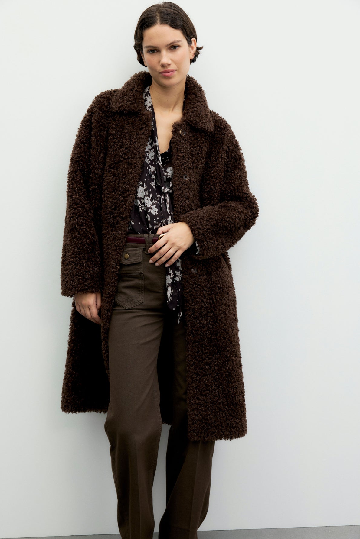 Woman standing against a plain wall wearing a long, dark brown manteau Teddy bear in tissu bouclé over a black and white floral blouse and brown pants, with short dark hair styled neatly.