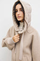 A woman wearing a beige doudoune, styled as a veste à capuche femme with a quilted hood and ribbed cuffs, stands against a plain white background, looking at the camera.