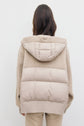 A person with long dark hair, seen from behind, is wearing a doudoune beige à capuche, featuring a quilted puffer section on the back and ribbed hem, paired with light pants.