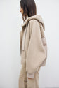 A person with long brown hair, wearing a veste à capuche femme in beige with knit cuffs and tan pants, stands sideways against a plain white background.