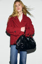 A person with long blonde hair wears a manteau rouge—an elegant red caban court—paired with blue jeans, holding a large black leather handbag, standing against a plain light background.