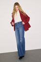 A person with long blonde hair wears a manteau rouge—a stylish red wool coat—over a white shirt, blue high-waisted wide-leg jeans, and black shoes, standing against a plain background.