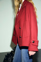 A person with long blonde hair wears a red manteau laine double-breasted coat over a white shirt and blue jeans, carrying a black handbag, against a plain background.