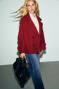 A person with long blonde hair wears a manteau rouge double-breasted coat, a light blouse, and blue jeans, and carries a black handbag while walking indoors against a plain background.