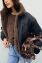 Woman in a brown sweater and black fausse fourrure parka courte, wearing light blue jeans with a brown belt, holding a brown leather handbag with gold hardware against a pale background.