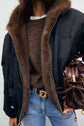 A person wearing a brown sweater, a black parka courte with fausse fourrure trim, blue jeans, and a brown belt, holding a brown handbag with a gold clasp. Their face is not visible.