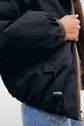 Close-up of a person wearing a black parka réversible with a zip and elastic cuff, paired with blue jeans. The person’s hand is visible near the jacket’s brown inner lining.