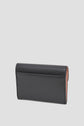 A black leather wallet, designed as a portefeuille super plat, is shown standing upright, closed, against a plain light gray background. The wallet has clean stitching and a minimal, modern design.