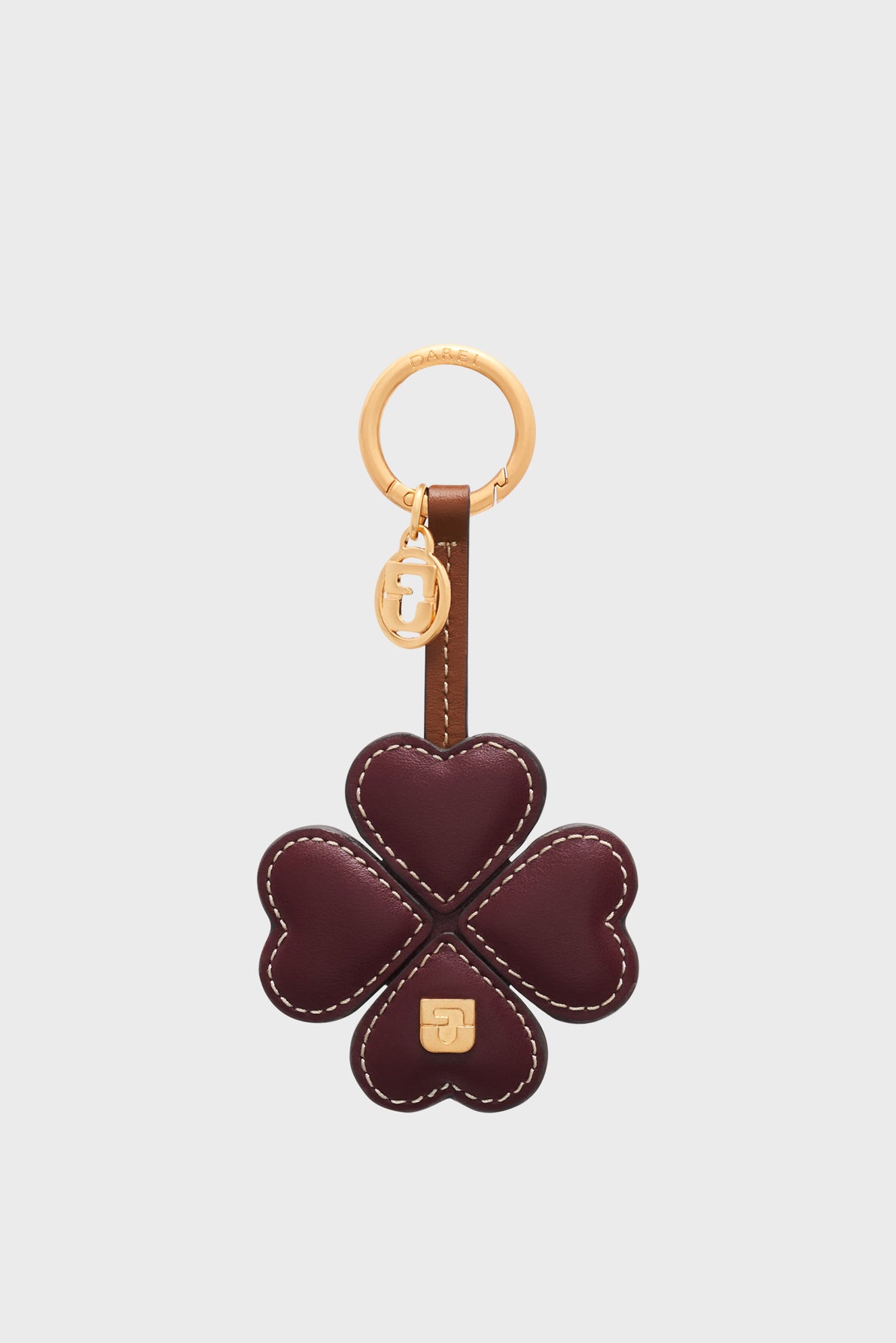A porte-clé trèfle featuring a gold ring and a dark red, four-leaf clover charm crafted from heart-shaped cuir de vachette petals, adorned with gold logo accents and detailed white stitching along the edges. This keychain is both a stylish accessory and a porte-bonheur to carry with you.