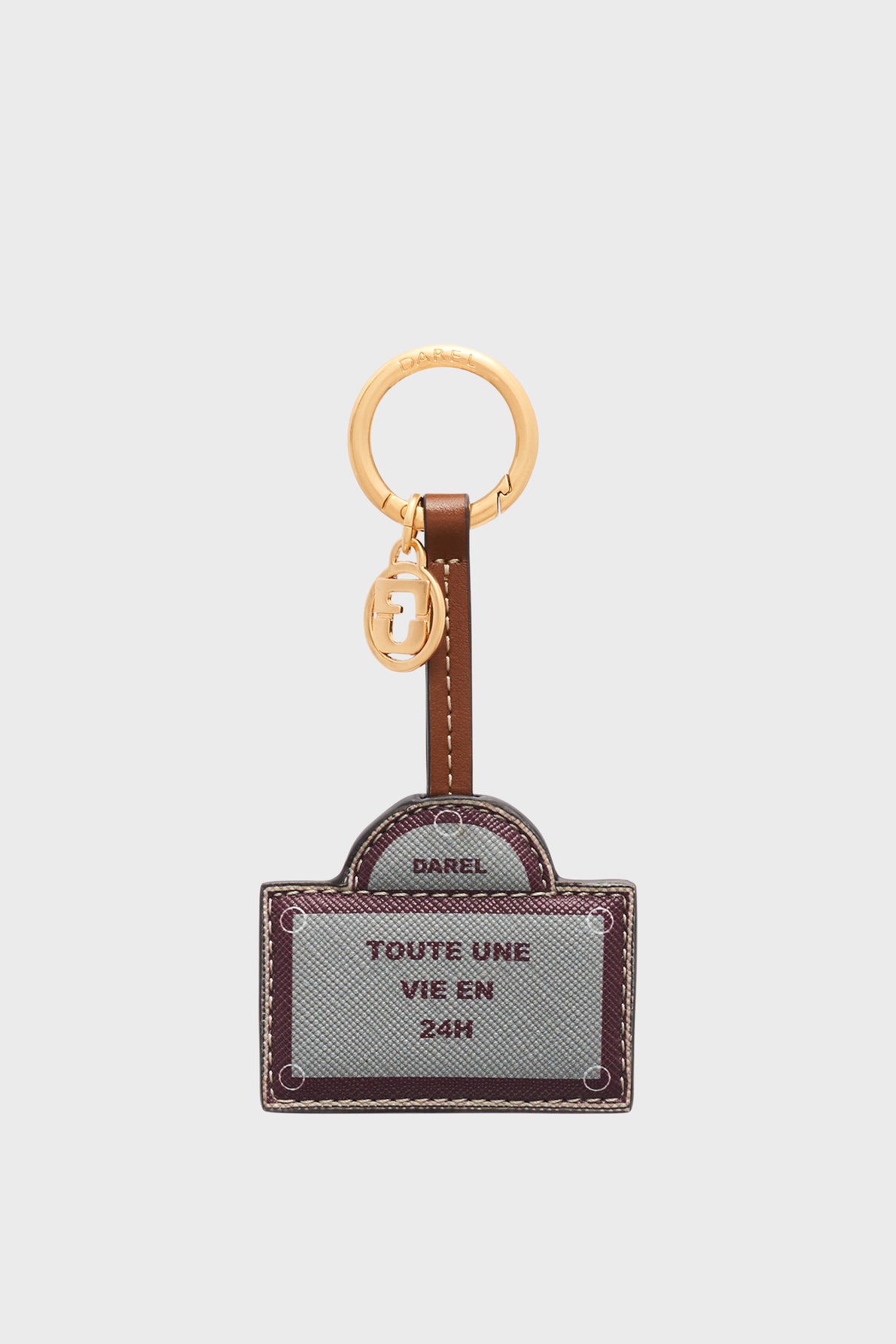 A gold keyring with a brown leather tag and a metal charm. The tag has gray and brown stitching and reads DAREL TOUTE UNE VIE EN 24H in uppercase letters. This élégant accessoire parisien is adorned with a médaille GD, adding a signature touch of style to your everyday essentials.