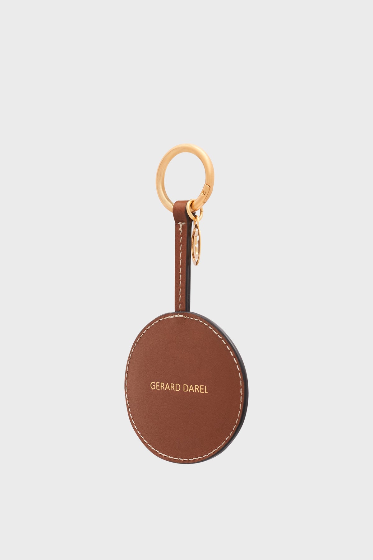 Round brown leather keychain with white stitching, featuring GERARD DAREL in gold letters, attached to a gold keyring with a small gold charm—radiating the élégance parisienne and allure charm of Gerard Darel—on a light gray background.
