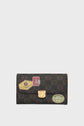 A dark brown patterned portefeuille Gerard Darel with a gold clasp, featuring travel-themed sticker designs including a Paris Eiffel Tower label and other decorative tags on the front.